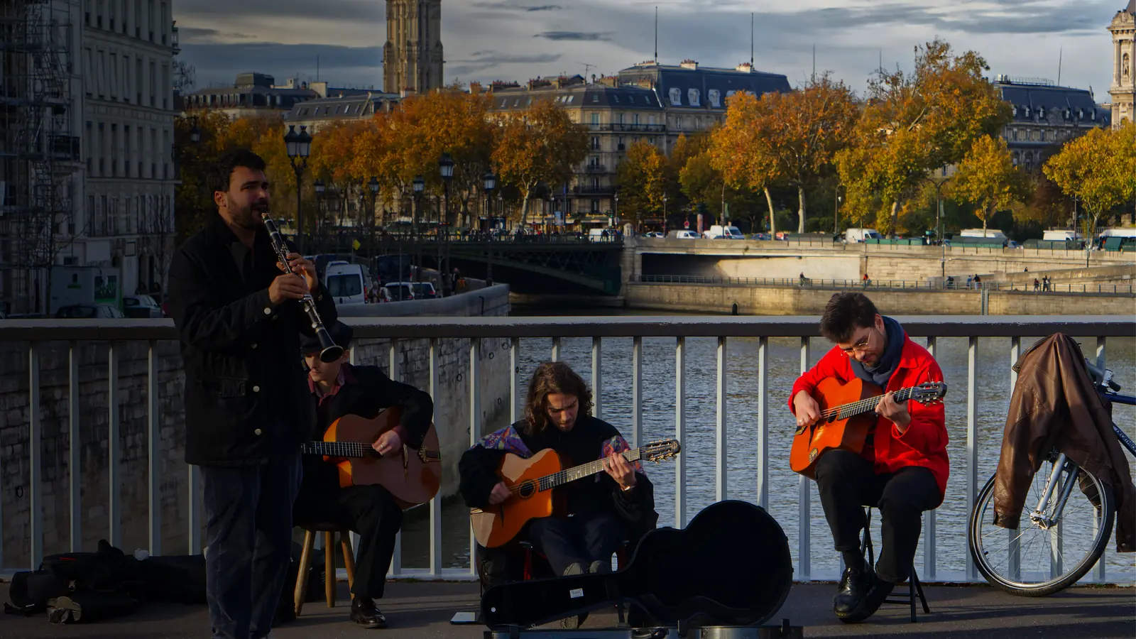 Four musicians playing on a bridge over a river in a city, instrument cases open at their feet
