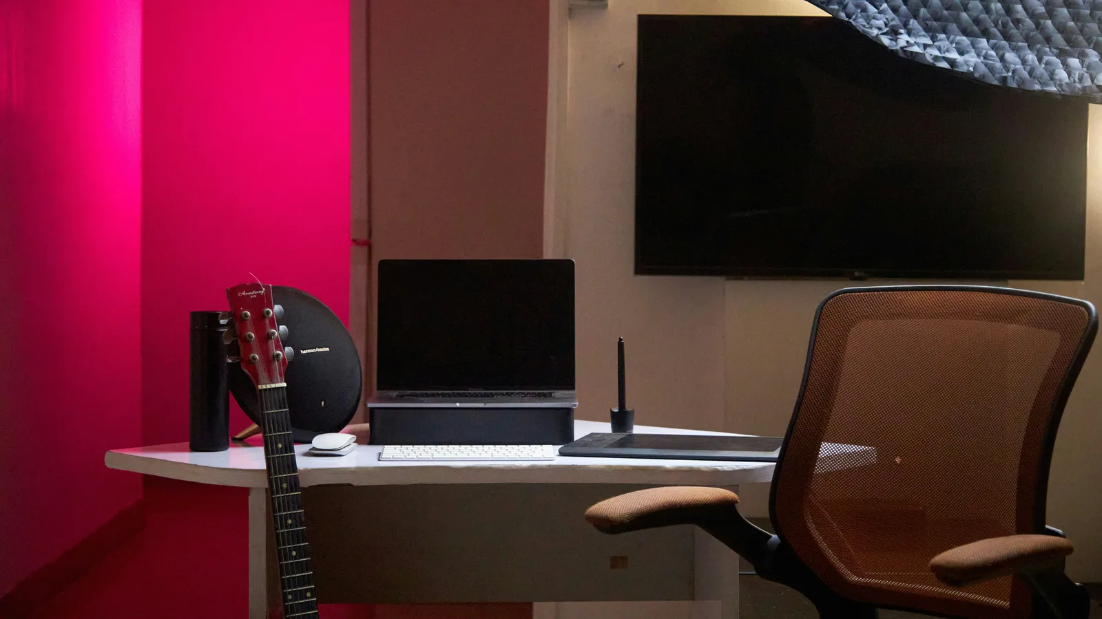 An empty studio workroom before anyone is in it: a desk with a laptop and keyboard, an acoustic guitar leaning against it, a mesh chair pulled back, magenta light on the wall