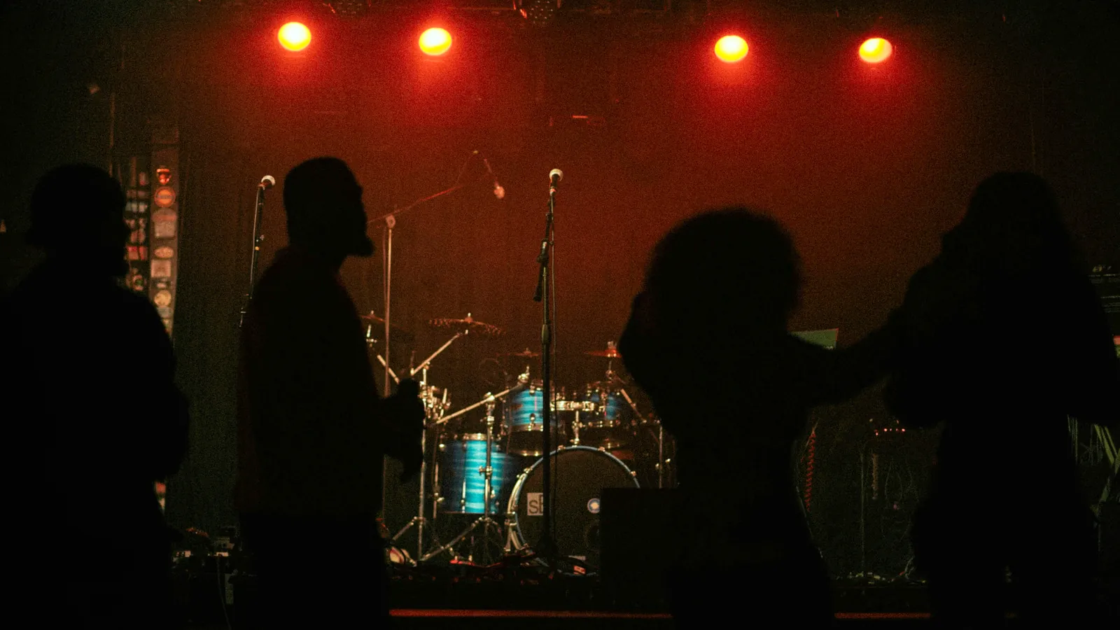 A small club stage under red lights, drum kit set up, a few silhouettes waiting