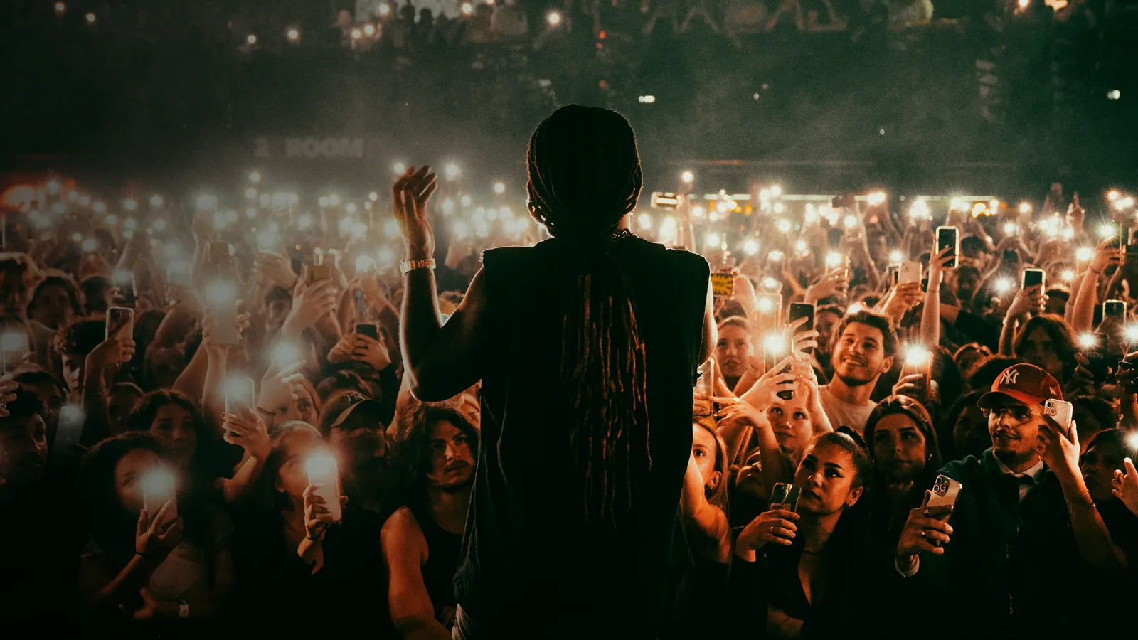 Seen from behind a performer on stage: a crowd filling the room, hundreds of phone lights held up
